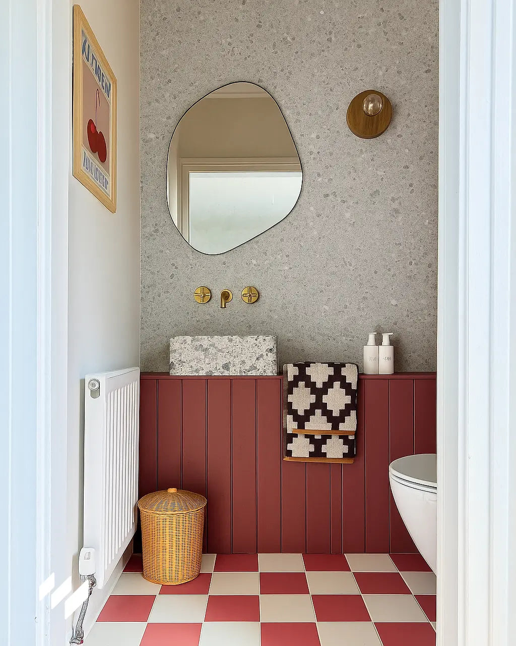 Aseo moderno con suelo de baldosas rojas y blancas, zócalo de listones burdeos, lavabo de piedra y pared trasera revestida con panel SPC efecto terrazo.