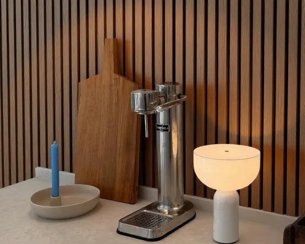 Rincón de cocina o coffee station con pared revestida con Revestimiento de Pared 3D Palillería Acústica en Roble Ahumado de Revestimientos Pared, aportando un toque contemporáneo. Encimera minimalista decorada con una vela azul en un cuenco de cerámica, una tabla de cortar de madera maciza y una lámpara de mesa blanca de diseño contemporáneo. En el centro destaca un grupo de extracción manual de acero inoxidable para café espresso