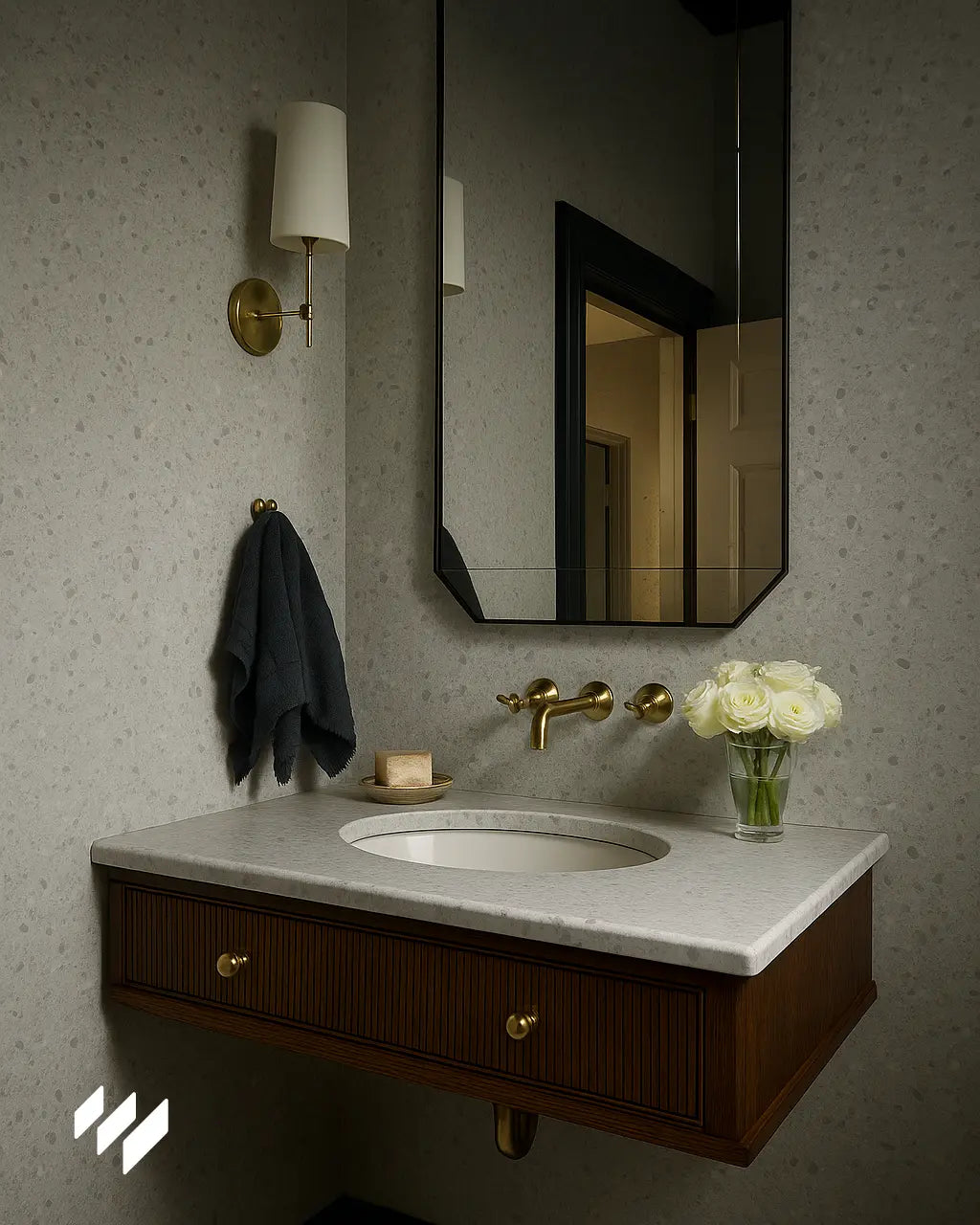 Lavabo de baño con grifería dorada y pared revestida con panel SPC efecto terrazo en tono gris claro, acompañado de espejo geométrico y aplique de pared elegante.