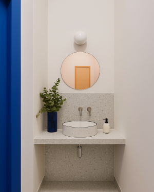Lavabo minimalista con encimera y pared trasera revestidas con panel SPC efecto terrazo, espejo redondo y grifo mural en un espacio compacto.