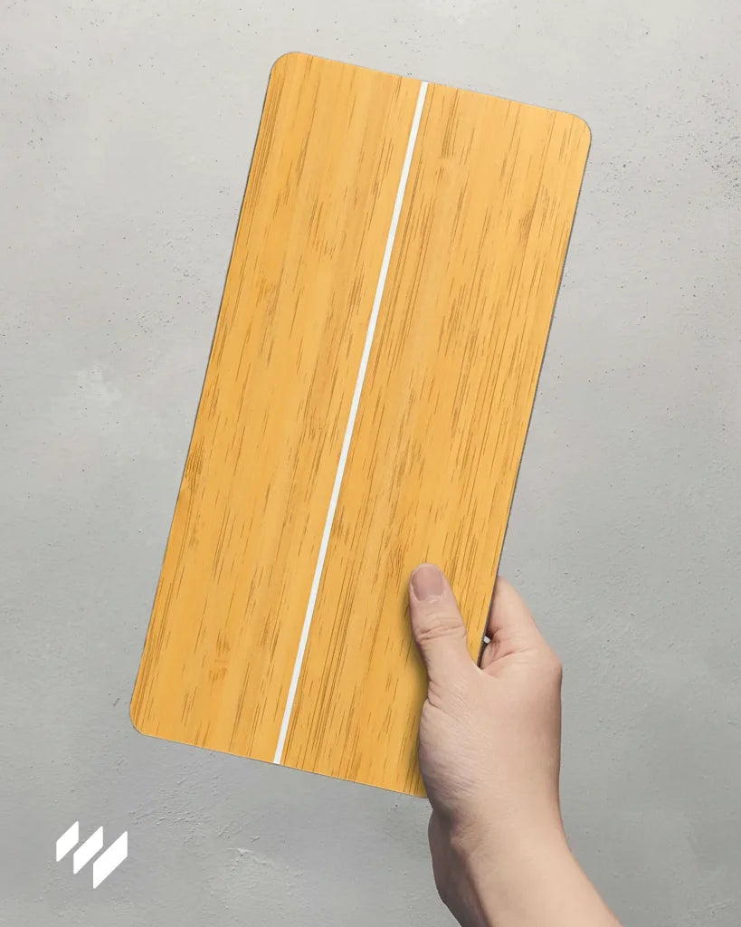 Mano sosteniendo muestra de Panel SPC efecto madera cerámica Versa, tono roble dorado con ranura blanca central, sobre fondo gris.