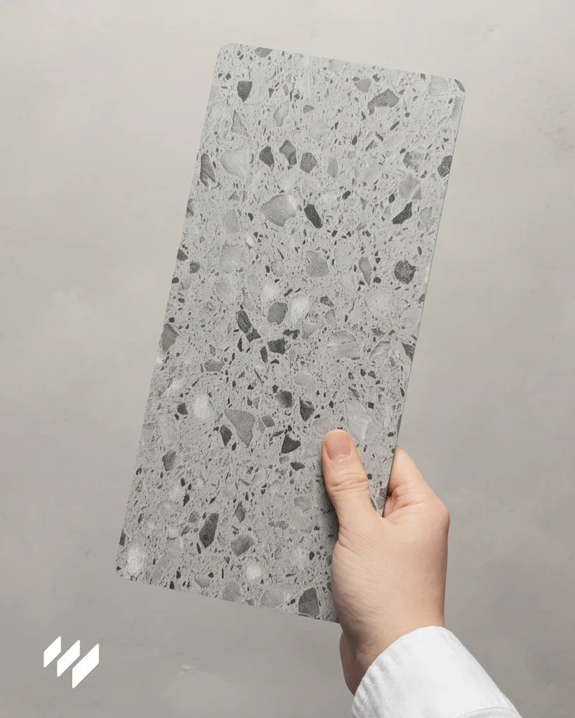 Muestra de revestimiento SPC para pared de ducha efecto terrazo gris oscuro Versa, sostenida con la mano sobre fondo neutro.