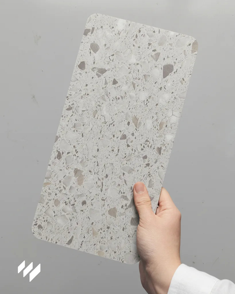 Mano sosteniendo muestra del revestimiento SPC efecto terrazo gris claro de Revestimientos Pared, ideal para baños modernos.