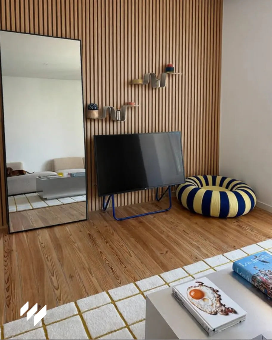 Salón moderno con pared de panel acústico roble natural de revestimientos pared, televisión apoyada, espejo de cuerpo entero y puff rayado.