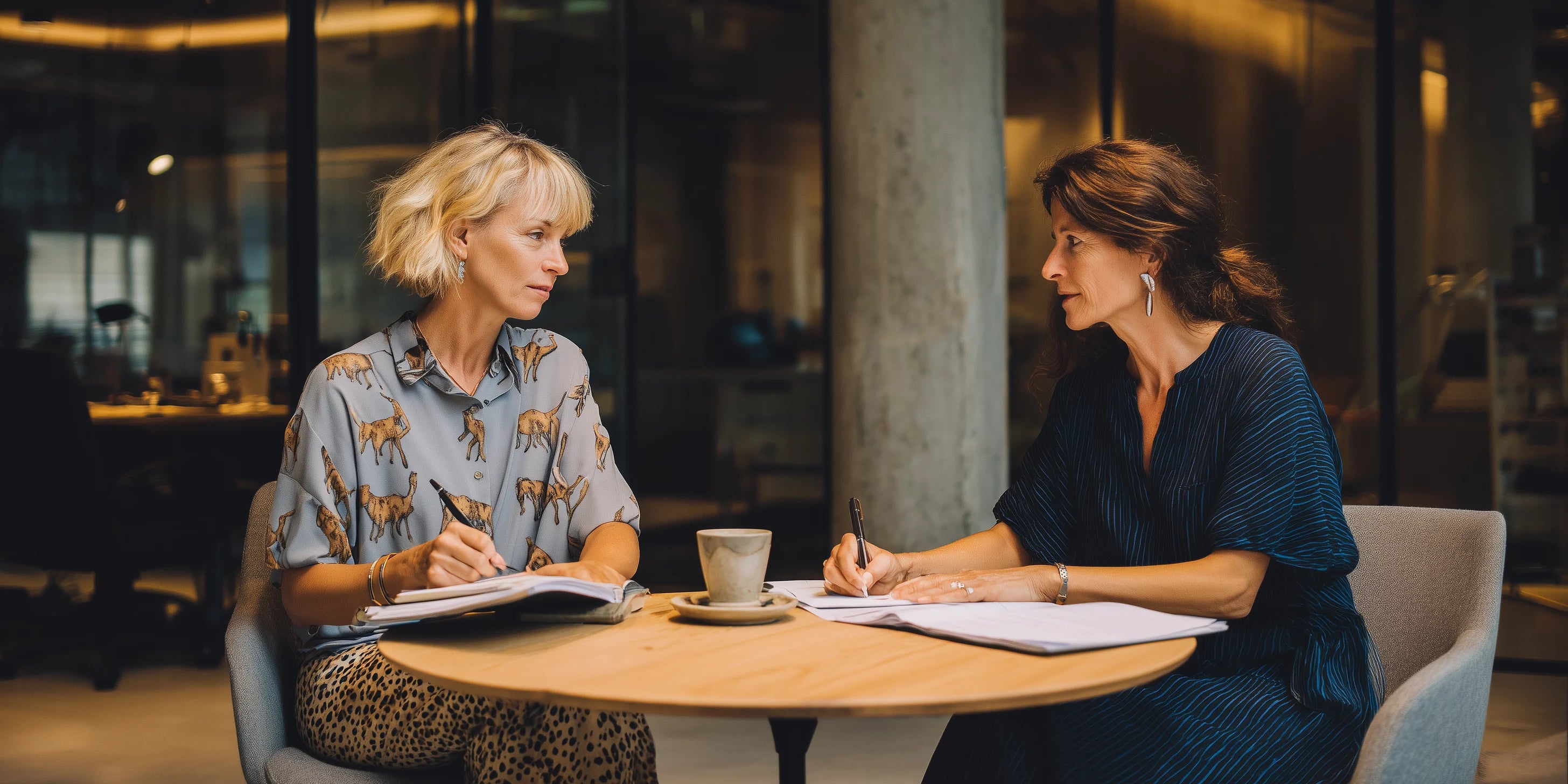 Reunión de dos mujeres tomando notas en una oficina moderna de Madrid, sentadas frente a una mesa redonda de madera, con ambiente profesional y acogedor.