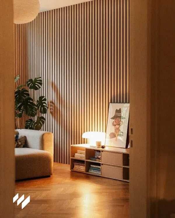 Pared decorada con paneles acústicos de listones en roble natural de revestimientos pared en un salón moderno con sofá curvo y lámpara de mesa.