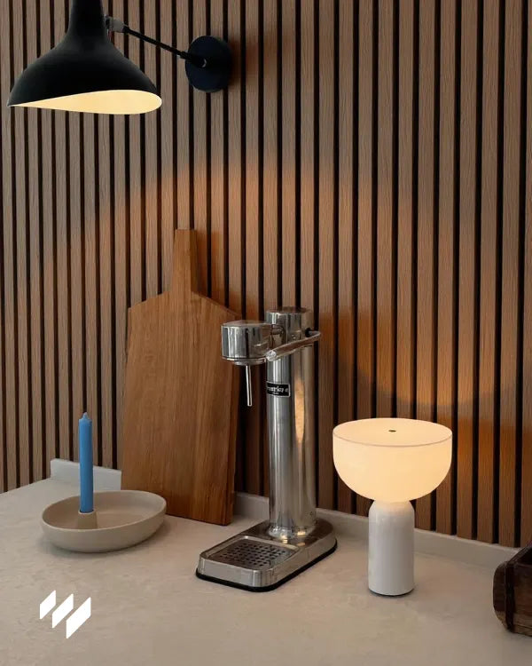 Rincón de cocina o coffee station con pared revestida con Revestimiento de Pared 3D Palillería Acústica en Roble Ahumado de Revestimientos Pared, aportando un toque contemporáneo. Encimera minimalista decorada con una vela azul en un cuenco de cerámica, una tabla de cortar de madera maciza y una lámpara de mesa blanca de diseño contemporáneo. En el centro destaca un grupo de extracción manual de acero inoxidable para café espresso