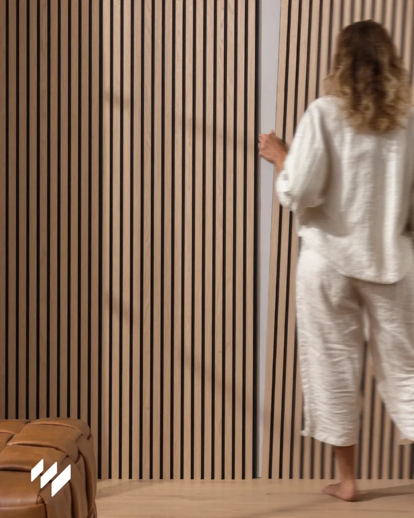 Mujer interactuando con un panel acústico de listones en color roble natural de revestimientos pared, instalado en una pared moderna con suelo de madera clara.