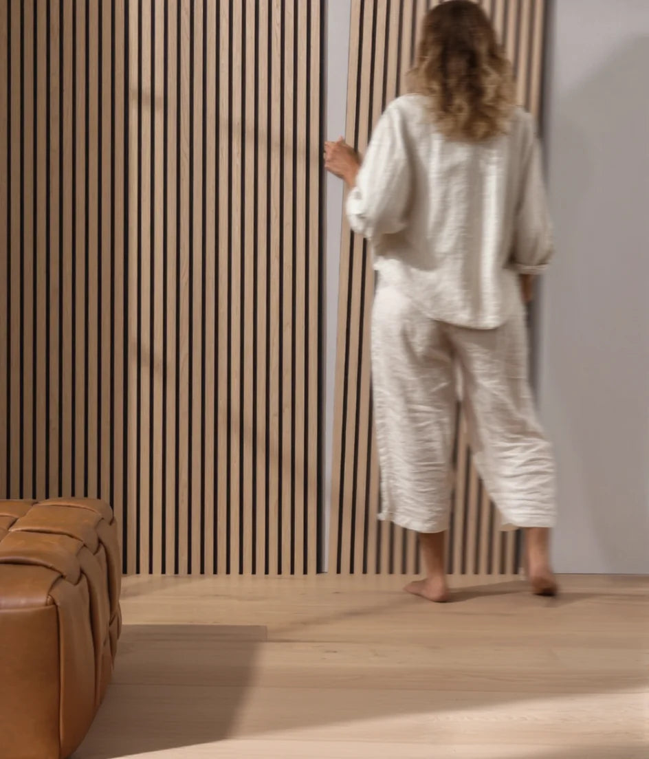 Mujer interactuando con un panel acústico de listones en color roble natural, instalado en una pared moderna con suelo de madera clara.
