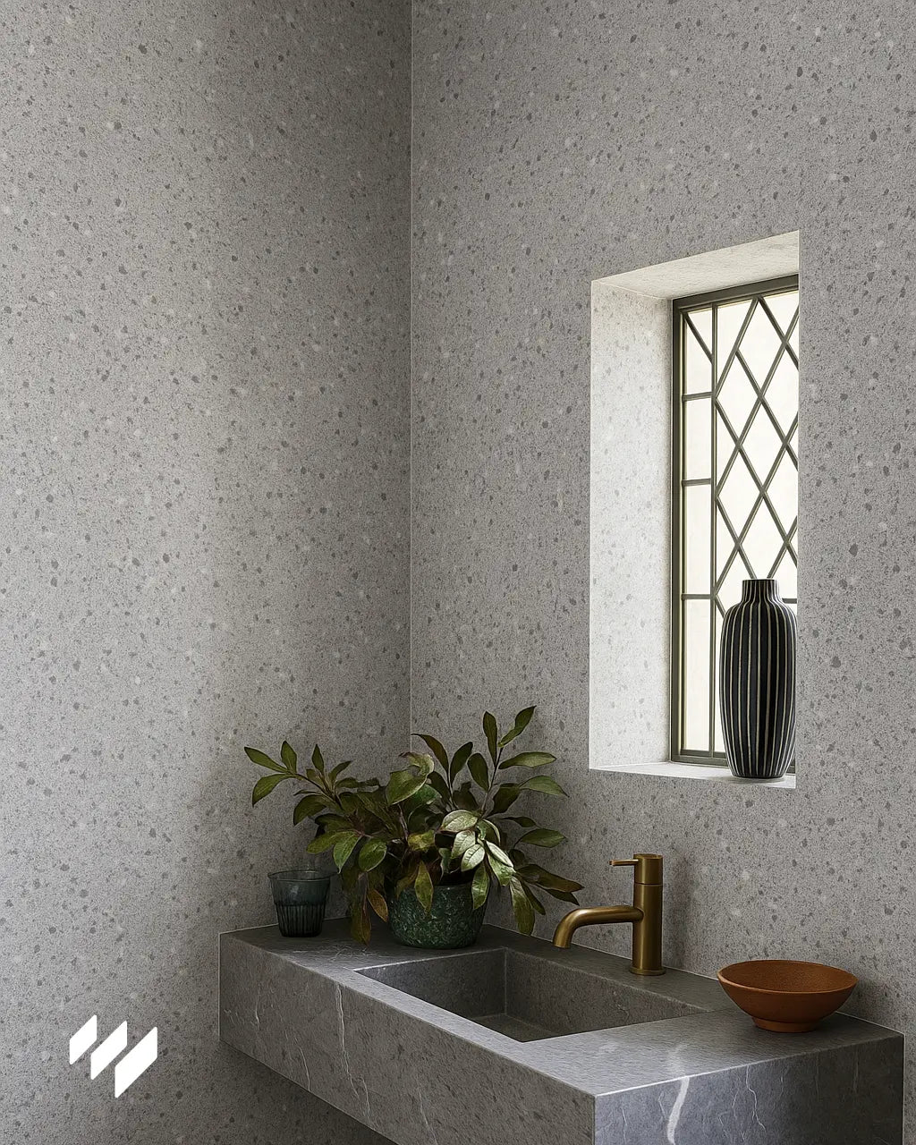  Lavabo de piedra gris con grifería dorada, plantas decorativas y fondo de pared revestido en SPC efecto terrazo en tono gris con ventana decorativa.

