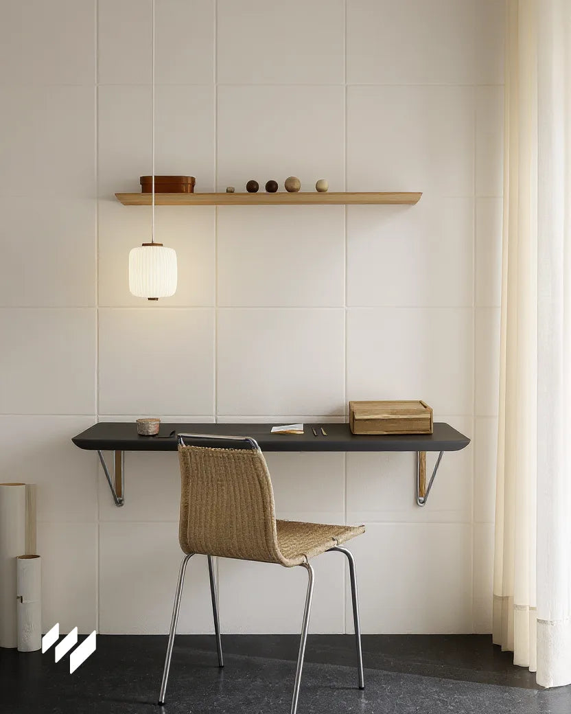 Espacio de estudio o escritura con diseño minimalista, decorado con paneles acústicos blancos de 50x50cm en la pared. Incluye un escritorio flotante negro, una silla de fibras naturales, una lámpara colgante cálida y una estantería de madera con objetos decorativos, junto a una cortina translúcida que deja pasar luz suave.