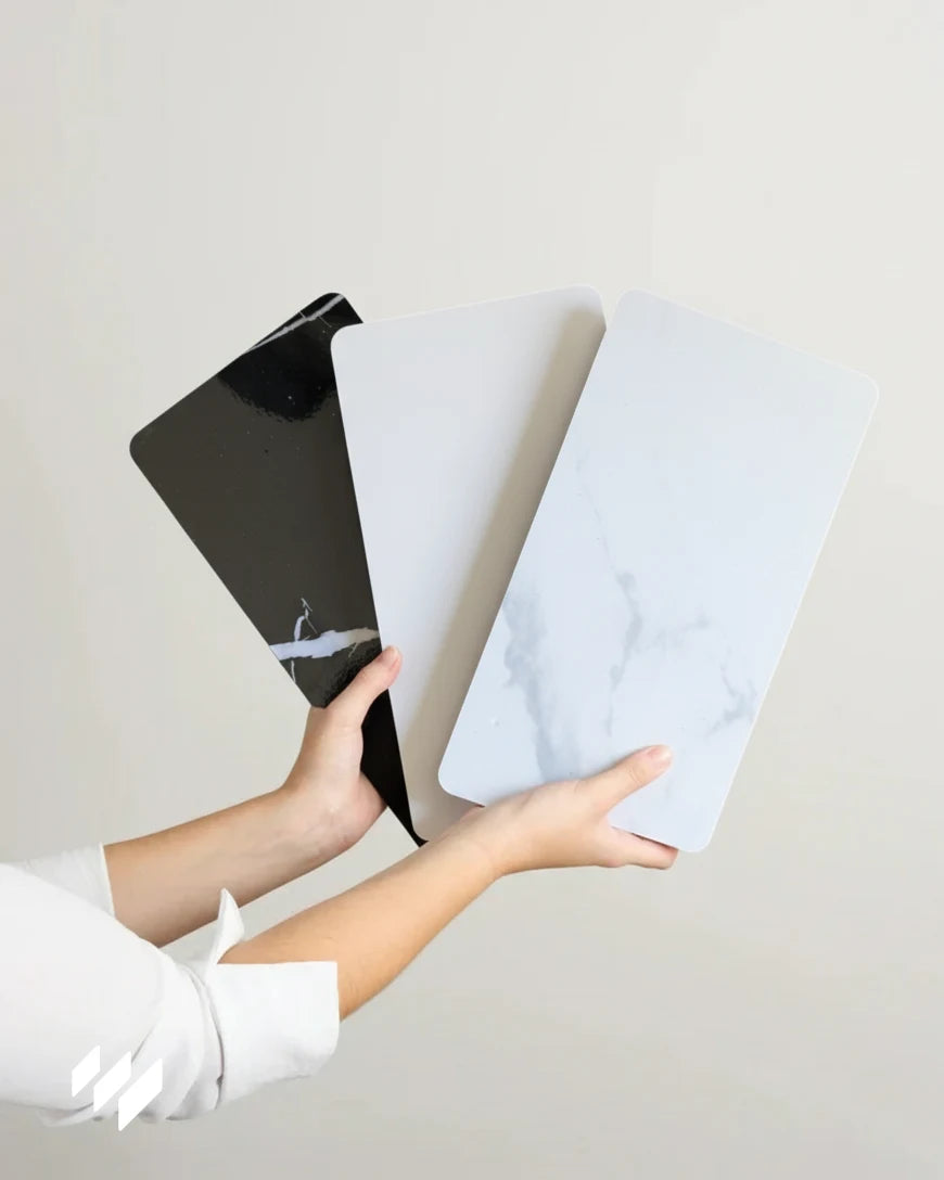 Manos sosteniendo tres muestras de paneles WPC efecto mármol de Revestimientos Pared: Negro Marquina, Calacatta Blanco alto brillo y Calacatta Blanco mate.