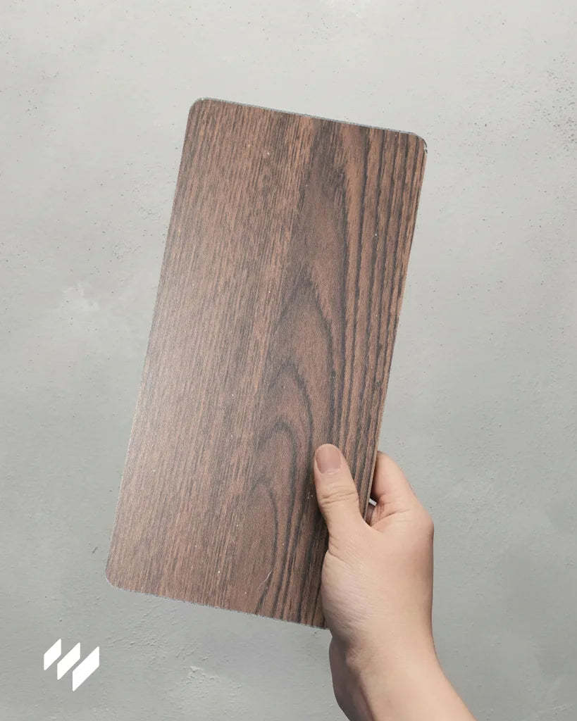 Muestra del panel decorativo WPC efecto madera Roble Nocturno sostenido con una mano sobre fondo gris — Revestimientos Pared.