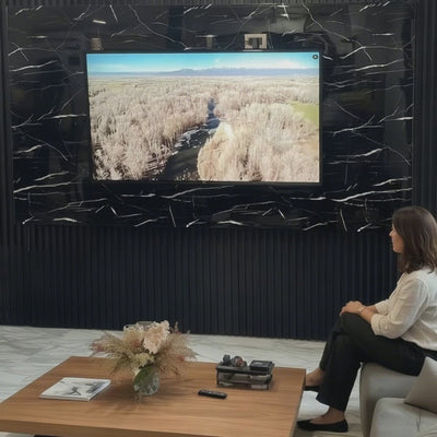 Marta, en su salón de Barcelona, observa un documental de naturaleza en la televisión montada sobre una pared de Panel WPC Alto Brillo efecto Mármol Negro Marquina, con fondo de listones negros y suelo marmoleado.