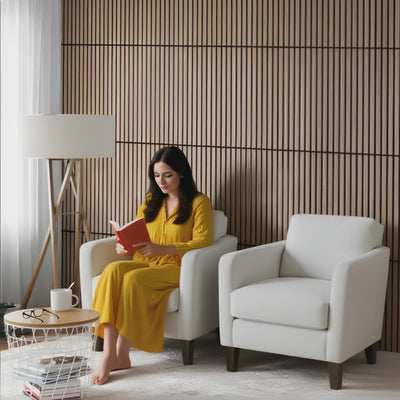 La mujer de cabello largo y oscuro, vestida con un conjunto holgado amarillo de tela suave, está sentada descalza en un sillón bajo. Lee un libro con expresión tranquila e introspectiva, con una postura relajada y una pierna ligeramente apoyada en el sillón. La luz se filtra suavemente por la ventana de la izquierda, proyectando sombras suaves.
