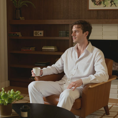 Un joven relajado y con estilo, de cabello corto y despeinado, vestido con una camisa de lino blanca semitransparente con rayas y un pantalón blanco holgado (de la imagen de la izquierda), está sentado de manera casual en un sillón de cuero color caramelo cálido en una sala de estar de estilo moderno de mediados de siglo (de la imagen de la derecha). Sostiene una taza de café con una mano apoyada en el brazo del sillón y la otra sujetando suavemente la taza. Su postura es relajada pero compuesta, con una pi